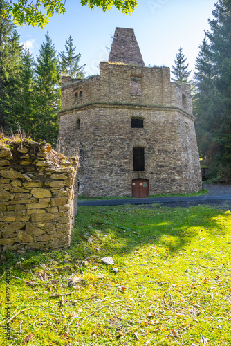 This former lime kiln near Kovarska in the Ore Mountains showcases stone architecture surrounded by lush greenery. Sunlight filters through trees, highlighting the historic structure's unique design. photo