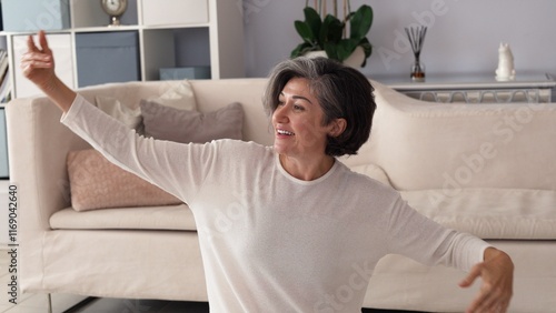 Принти на полотні Caucasian woman, gray streaked hair, white sweater, arms gracefully lifted, smiling softly in serene living space, expressing peace, calm, joyful presence, enjoying relaxed moment indoors
