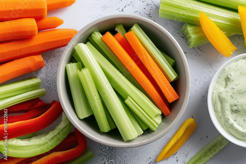 A close-up of freshly cut celery and carrot sticks arranged in a bowl with colorful bell peppers and a side of creamy green dip on a white table.
 photo