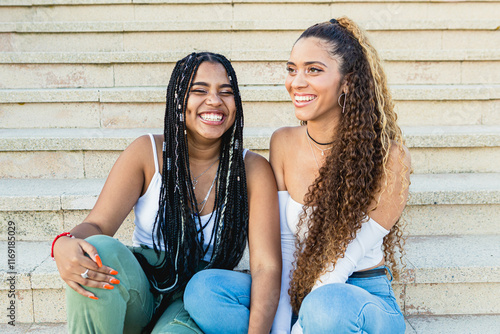 Two young latin women laughing together sitting on the stairs of a building