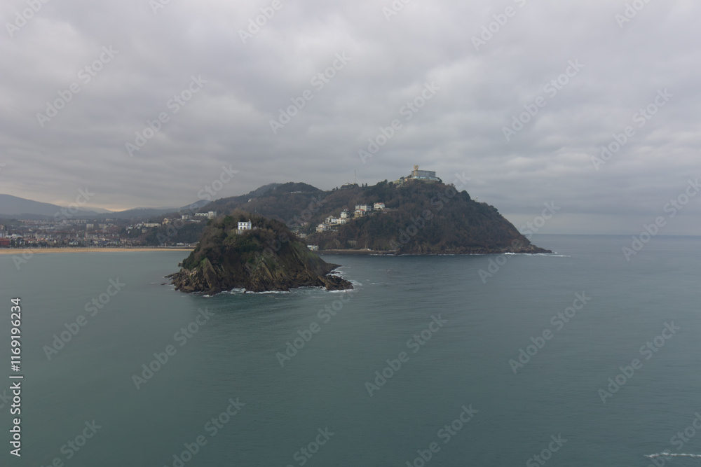 Scenic view of Santa Clara Island and Monte Igueldo with turquoise waters and coastal cityscape under a cloudy sky in San Sebastian, Spain. Scenic view of Santa Clara Island and Monte Igueldo with turquoise waters and coastal cityscape under a cloudy sky in San Sebastian, Spain.