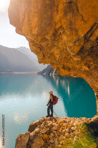 The front view of a woman stands on a cliff by Tientang Hu lake, Xinjiang, China. The curved stone corner of the lakeside walkway. The sun was rising with yellow light reflected on the water.