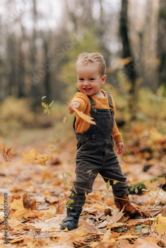 A little boy in a yellow sweater stands in a park with leaves