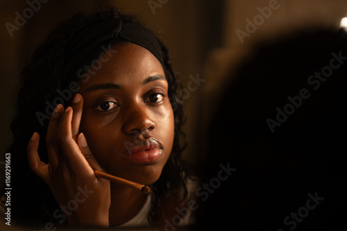Young woman carefully applying makeup, holding brush and gazing intently into mirror within shadowy interior, styling beauty routine