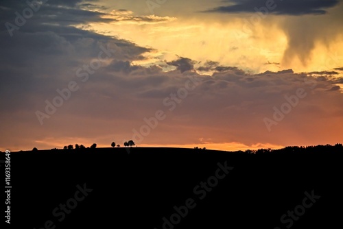 Wallpaper Mural Dramatic evening sky with layers of dark clouds, illuminated by warm orange and yellow hues. A line of silhouetted trees atop a distant hill adds depth to this striking natural scene. Torontodigital.ca