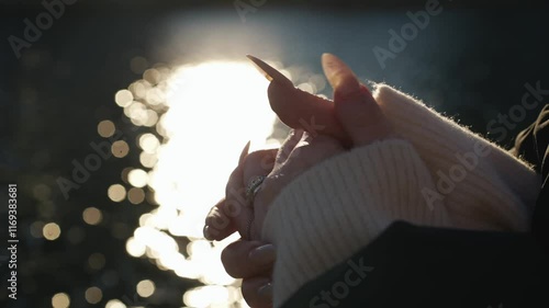 a girl with long nails on her hands stands on the river bank on a sunny day.
The glare of the sun on the water and women's hands in a warm sweater.
A girl strokes her hands while standing on the emban photo