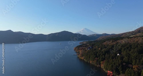 Forward tracking aerial footage of Mount Fuji rising over Lake Ashi, surrounded by forested hills and a red torii gate on the shoreline, under a clear blue sky.