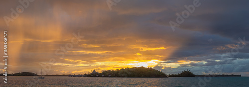 Obraz na płótnie A serene and picturesque scene of Bora Bora at sunset, a sailboat is anchored in the calm ocean waters, with the lush, green island and its mountainous terrain in the background. w salonie