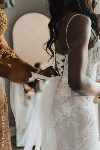 Mother adjusting and tying the intricate lace detailing of a bridal gown, capturing a tender wedding preparation moment with floral accents and soft natural light