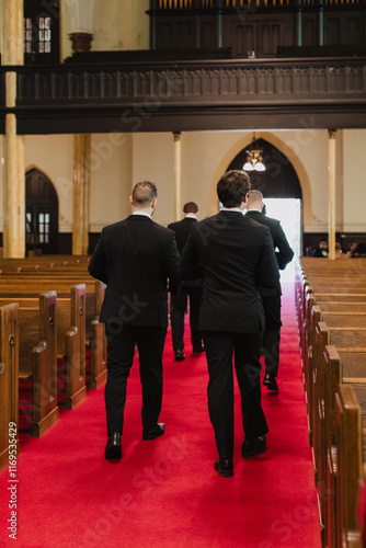 Formal procession inside a historic church with red carpet aisle, featuring groomsmen in black suits walking toward the altar under ornate woodwork and stained glass windows