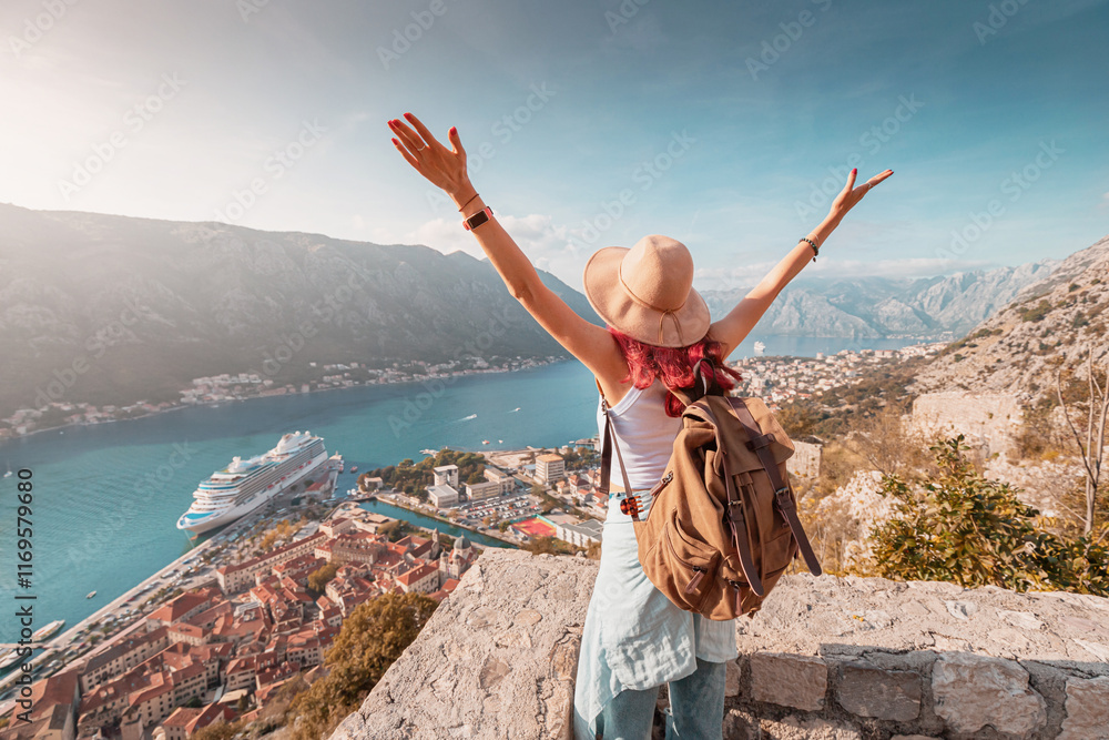 Female tourist with backpack and hat raising arms, celebrating scenic view of Kotor bay from mountain viewpoint at fortress in Montenegro, with cruise ship docked in harbor below