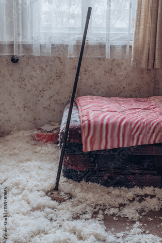 Bed with scattered feathers and a cleaning tool in an untidy room filled with sunlight photo