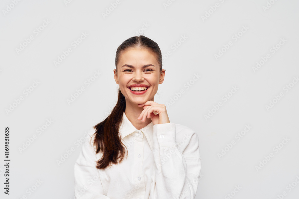Smiling woman with long brown hair, wearing a white shirt against a plain light gray background, expressing confidence and happiness with a friendly gesture Smiling woman with long brown hair, wearing a white shirt against a plain light gray background, expressing confidence and happiness with a friendly gesture