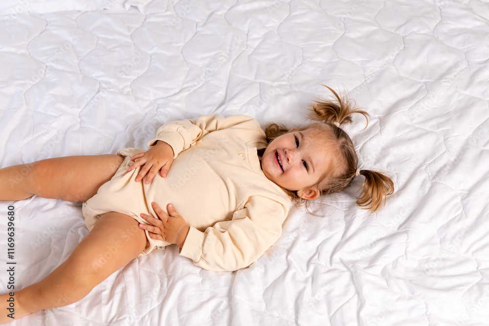 Naklejka premium Happy baby in bed. Cute little newborn girl in bodysuit lying on bed in bedroom. Infant baby resting, playing on white blanket at home. Motherhood and childcare concept