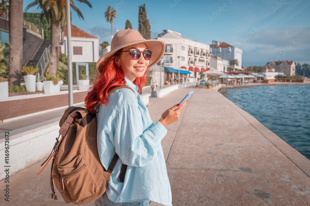 Happy red haired woman using smartphone while walking on seafront promenade near hotels and restaurants