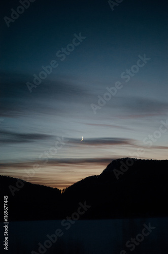 Moonlit crescent over silhouetted mountain ridge under a serene teal sky with wispy clouds during a winter evening..Europe, Norway, 02.01.2025  [The photographer confirms the authenticity of the photo