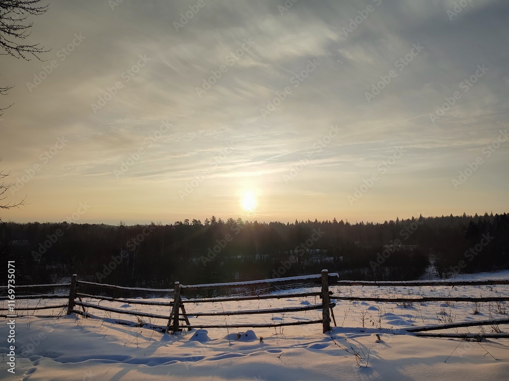 beautiful view of the rising sun in the sky in the clouds, against the background of a dark forest and half a mile covered with bright white snow behind an old, dilapidated, rural fence on a winter beautiful view of the rising sun in the sky in the clouds, against the background of a dark forest and half a mile covered with bright white snow behind an old, dilapidated, rural fence on a winter