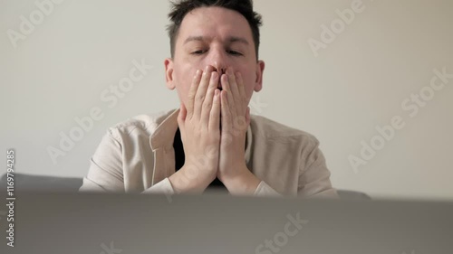 Young man feeling stressed while working on laptop in a modern home interior. Disappointed man covering face palm thinking. Depressed businessman looking frustrated.