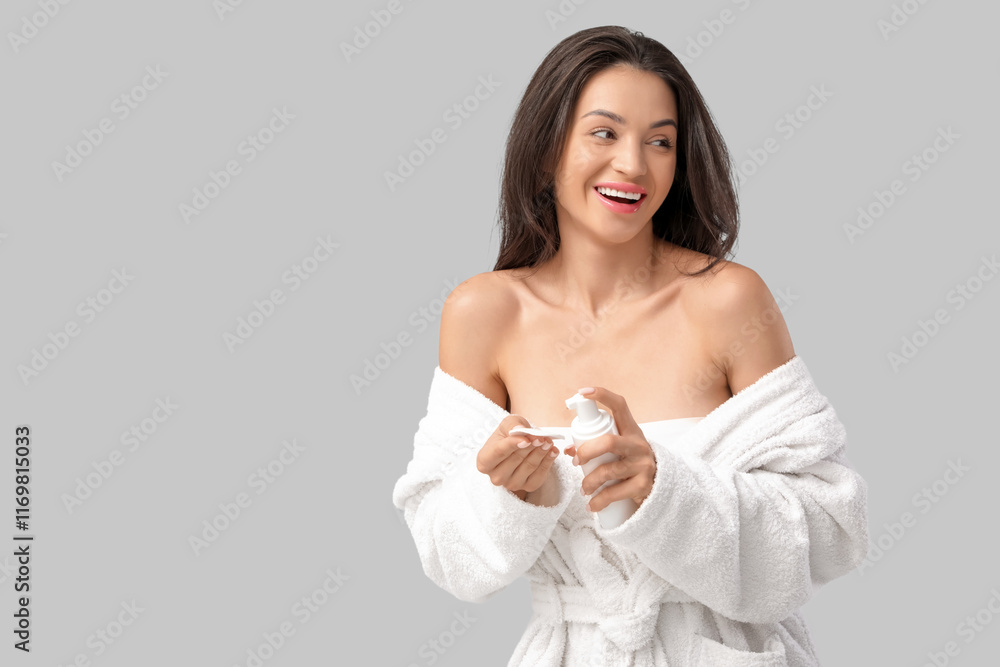 Beautiful young happy woman in bathrobe with cotton pad and bottle of cosmetic product on grey background Beautiful young happy woman in bathrobe with cotton pad and bottle of cosmetic product on grey background