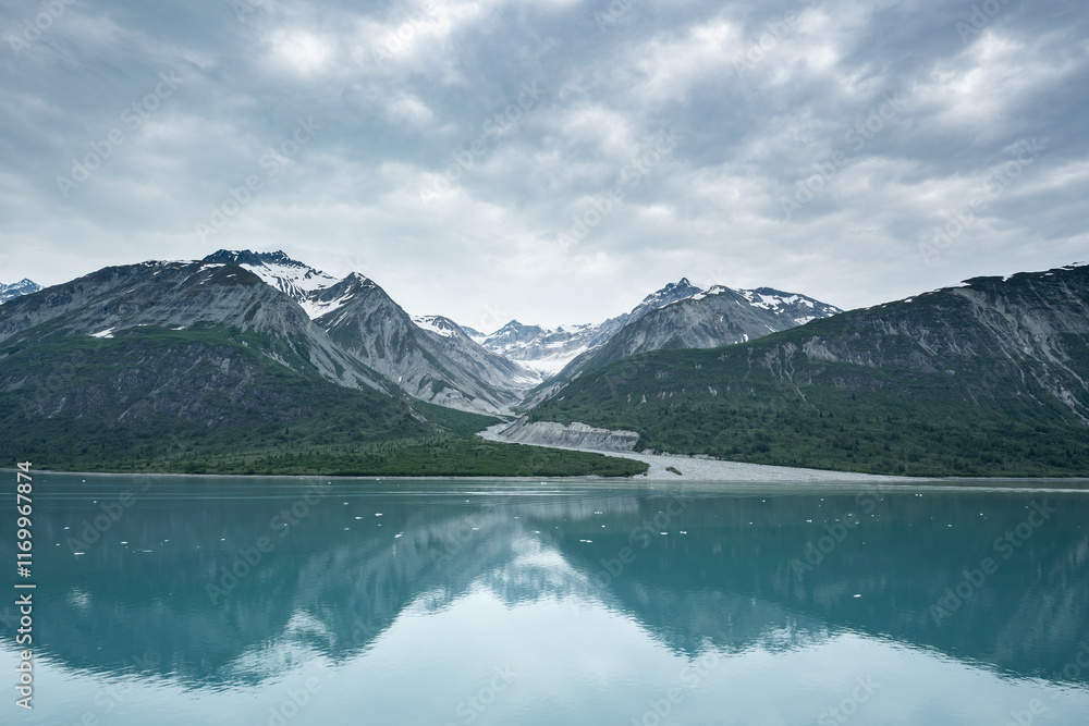 Fototapeta premium Beautiful snowy mountain peaks rising up from the shoreline of a calm body of water with a stunning reflection below and a cloudy sky overhead. 