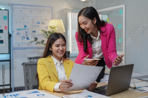 Two happy, smiling businesswomen collaborating and discussing a project while working with printed documents and a laptop in a modern office environment