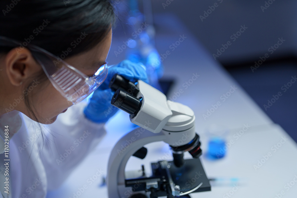 Female scientist wearing lab coat and safety glasses using microscope doing scientific research in modern laboratory, developing new treatment Female scientist wearing lab coat and safety glasses using microscope doing scientific research in modern laboratory, developing new treatment