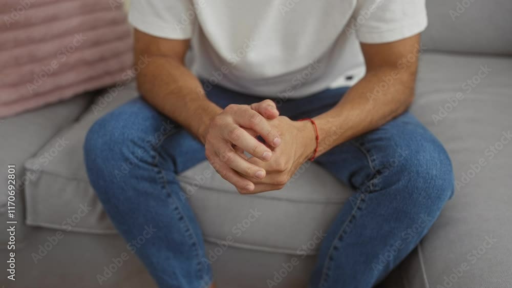 Young hispanic man sitting on a couch in a living room, with clasped hands and jeans, inside an apartment indoors Young hispanic man sitting on a couch in a living room, with clasped hands and jeans, inside an apartment indoors