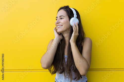 Isolated portrait of a beautiful young woman smiling and listening to the music with white headphones in front of a yellow wall