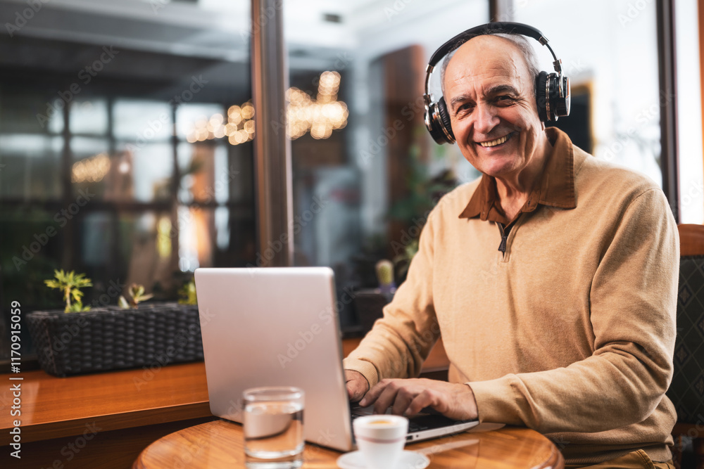 Fototapeta premium Smiling senior man in casual clothing with wireless headphones using laptop and drinking coffee while sitting in the cafe.