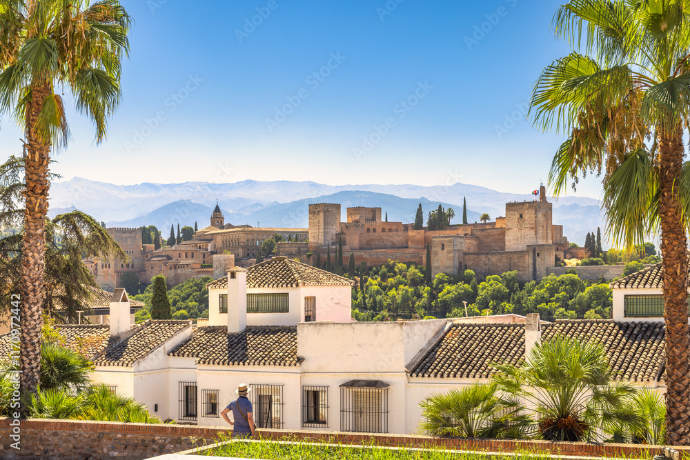 The Alhambra palace from viewpoint in Granada town in Spain. Historic castle viewed from a sun-drenched hillside, palm trees framing the scene. A person observes the view. The Alhambra palace from viewpoint in Granada town in Spain. Historic castle viewed from a sun-drenched hillside, palm trees framing the scene. A person observes the view.