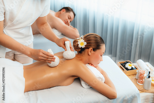 Hot herbal ball spa massage body treatment, masseur gently compresses herb bag on couple customer body. Tranquil and serenity of aromatherapy recreation in day lighting ambient at spa salon. Quiescent photo