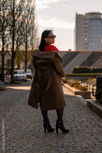 A confident woman in a stylish red blouse and black skirt posing outdoors in a modern urban setting during golden hour.