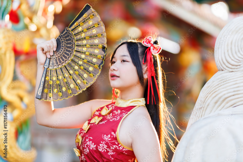 Portrait beautiful cute smiles Asian young woman wearing red traditional Chinese cheongsam decoration and holding a Chinese Fanning for Chinese New Year Festival at Chinese shrine in Thailand