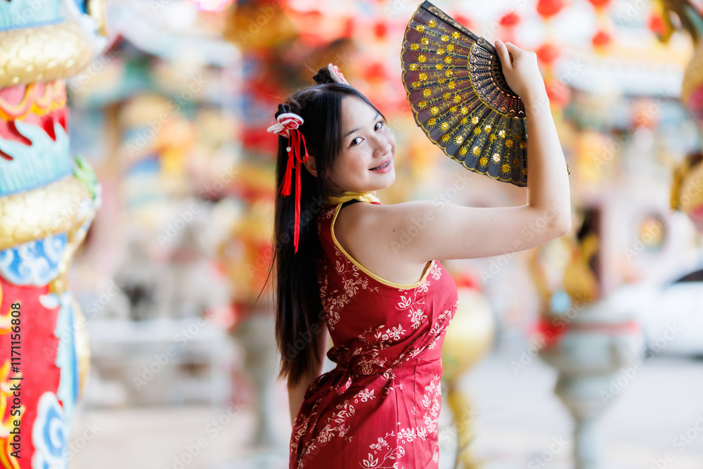 Portrait beautiful cute smiles Asian young woman wearing red traditional Chinese cheongsam decoration and holding a Chinese Fanning for Chinese New Year Festival at Chinese shrine in Thailand