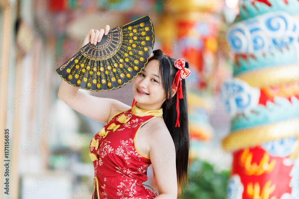 Portrait beautiful cute smiles Asian young woman wearing red traditional Chinese cheongsam decoration and holding a Chinese Fanning for Chinese New Year Festival at Chinese shrine in Thailand