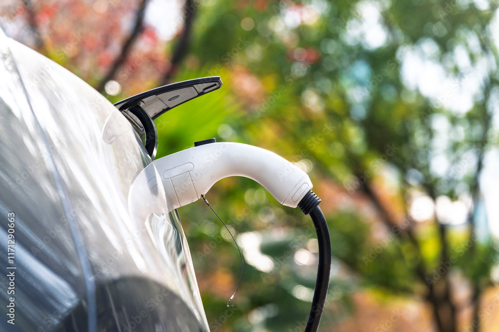 Closeup EV Car charger handle plugged in recharging EV car battery from EV charging station with green nature background. Eco-friendly alternative energy concept