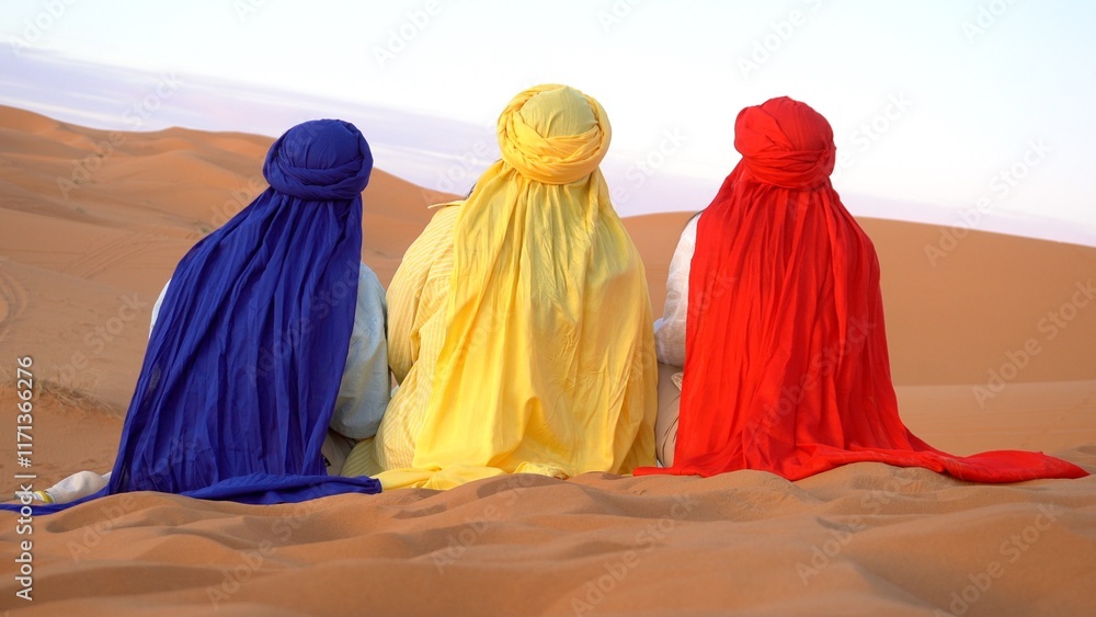 three islamic tourist arab women with colorful red, yellow and blue turbans sit in the Sahara desert in Morocco Africa watching the sand dunes and sunset - trip to Merzouga to visit African nature
