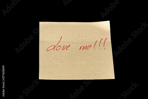 Yellow adhesive paper, similar to a Post-it note, on a black background with the words “Love MeE and three exclamation marks handwritten in red ink.