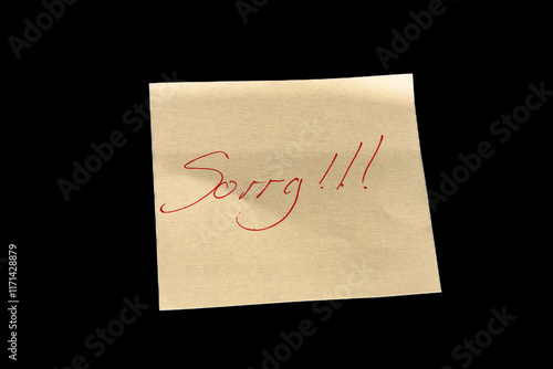 Yellow adhesive paper, similar to a Post-it note, on a black background with the word “Sorry” and three exclamation marks handwritten in red ink.