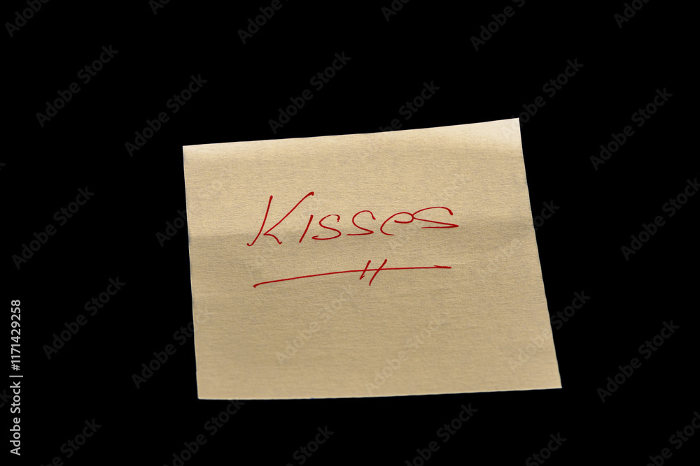 Yellow adhesive paper, similar to a Post-it note, on a black background with the word “Kisses” handwritten in red ink. Yellow adhesive paper, similar to a Post-it note, on a black background with the word “Kisses” handwritten in red ink.