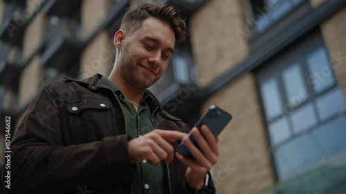 Low-angle view of concentrated young male navigating digital content, networking through mobile device. Close-up of cheerful man checking smartphone standing on urban street. Shooting in slow motion.
