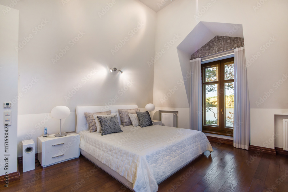 Fototapeta premium The interior of a bedroom in a luxurious country mansion with a beautiful view of the .lake from the window. White room decor, parquet floor.