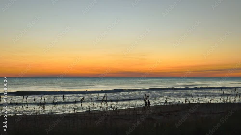 A couple, man and woman, walk along the beach during a breathtakingly beautiful sunrise sky with vibrant colors sun rising over the Atlantic Ocean A couple, man and woman, walk along the beach during a breathtakingly beautiful sunrise sky with vibrant colors sun rising over the Atlantic Ocean