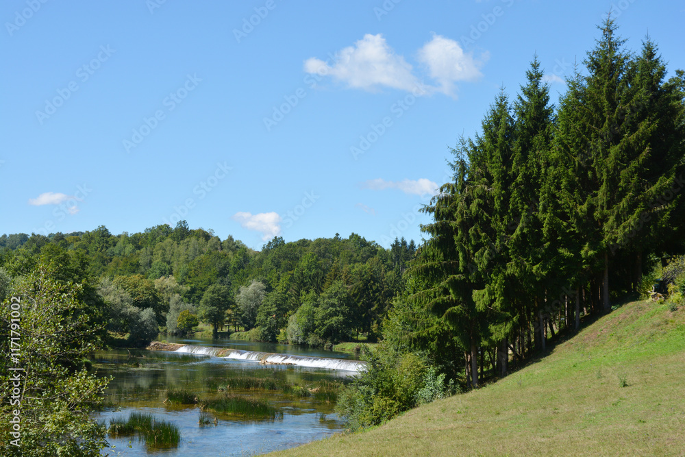 A picturesque view of the Dobra River near Jaškovo, featuring calm waters reflecting the blue sky and white clouds, a small dam creating a gentle waterfall, and lush green vegetation lining the banks A picturesque view of the Dobra River near Jaškovo, featuring calm waters reflecting the blue sky and white clouds, a small dam creating a gentle waterfall, and lush green vegetation lining the banks