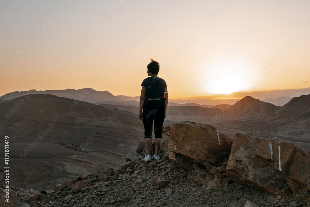 Woman stands alone on the high rock during sunset and looking at setting sun in distance Woman stands alone on the high rock during sunset and looking at setting sun in distance