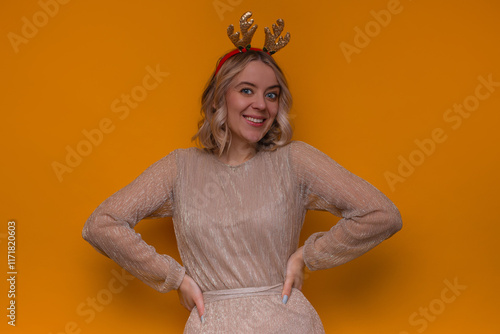 A cheerful woman wearing a sparkling gold dress and glittery reindeer antlers poses confidently against a vibrant orange background, embodying festive elegance and holiday joy. photo