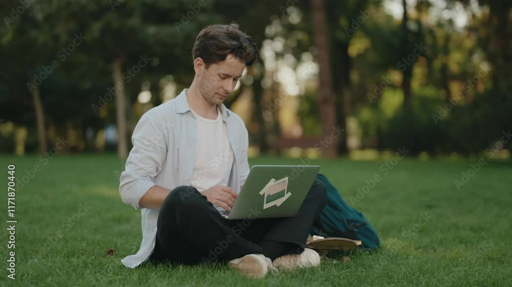 A young man sits in the grass in a park, working on his laptop. He appears focused and productive while working remotely. A young man sits in the grass in a park, working on his laptop. He appears focused and productive while working remotely.