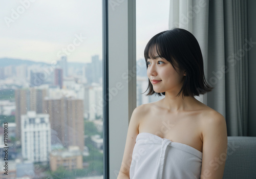 Asian lady after her morning facial skincare and beauty routine looking refreshed, wearing a white towel, relaxing near window view of city skyline outside her apartment. Free Download Asian lady after her morning facial skincare and beauty routine looking refreshed, wearing a white towel, relaxing near window view of city skyline outside her apartment. - GFXLoad