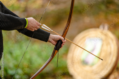 Archer's hand with wooden bow and arrow before shooting with an outdoor target in the background.