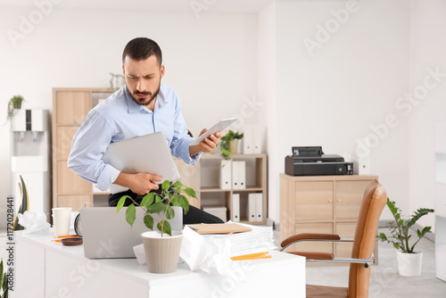 Busy male employee multitasking with laptop and tablet in a disorganized offi...